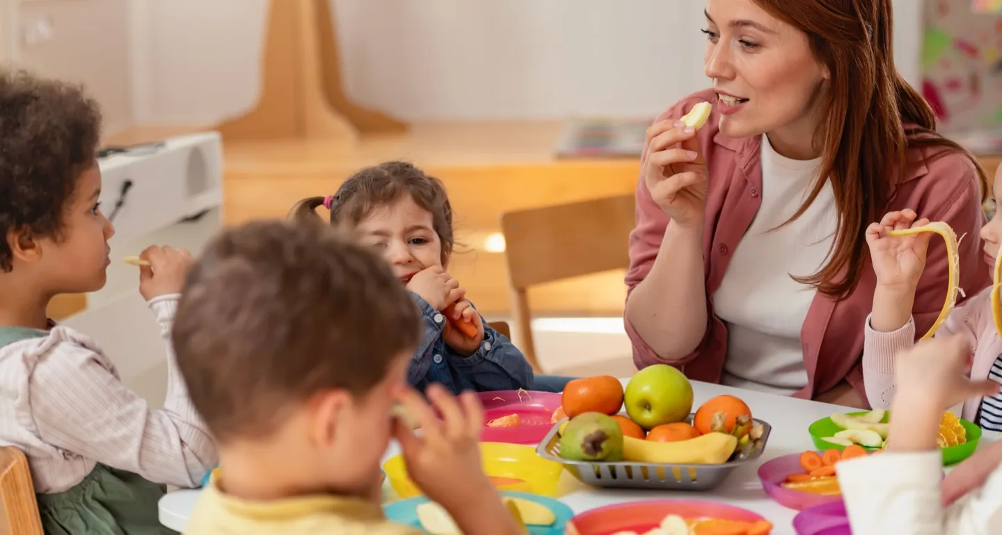 A female teacher sits with young students at a classroom table sharing snacks free from the Big Nine allergens. 