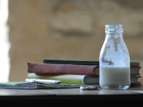 A small bottle of milk on a table beside textbooks and pencils