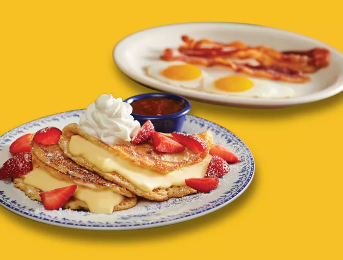 foreground: stuffed cheesecake pancake topped with whipped cream and strawberries on plate. background: two eggs, sunny-side-up and strips of bacon on plate