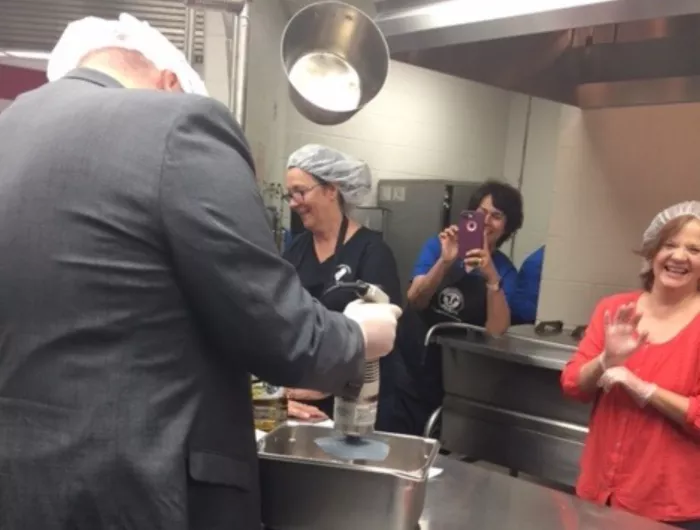 Staff in a school kitchen--several watch while one cooks