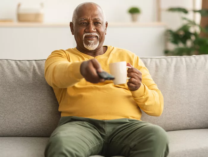 older man holding coffee and pointing remote towards the tv while sitting on a sofa