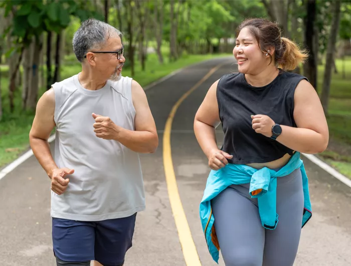 Man and woman with larger bodies unning on a paved trail in a tree lined park