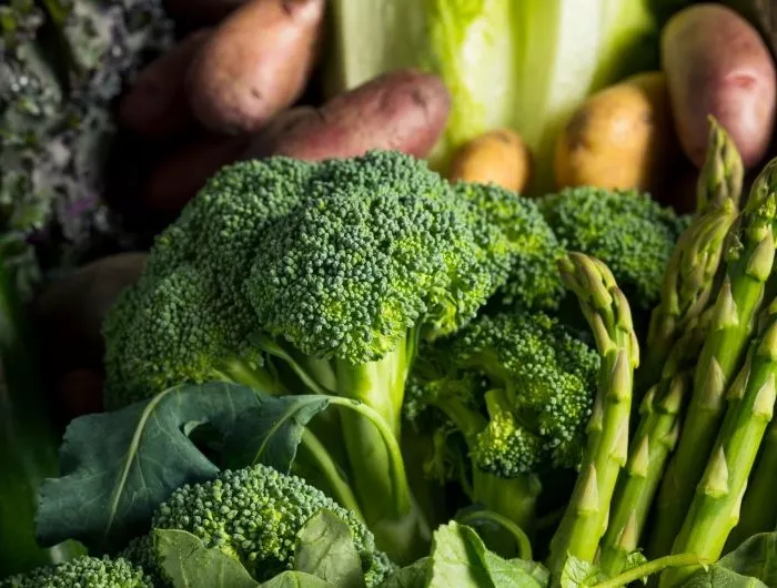 Seasonal produce - a closeup of broccoli and asparagus in the foreground, with potatoes, cabbage, collard greens and kale in the background.