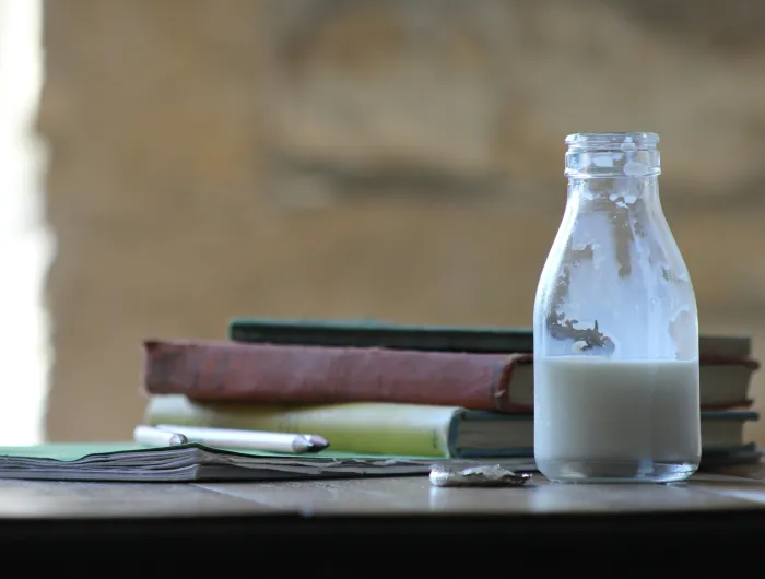 A small bottle of milk on a table beside textbooks and pencils
