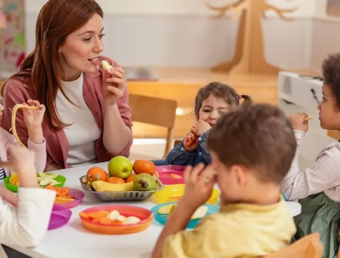 A female teacher sits with young students at a classroom table sharing snacks free from the Big Nine allergens. 