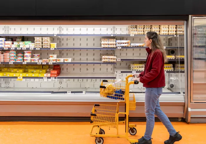 A woman pushes a cart past a nearly empty grocery store refrigerator section
