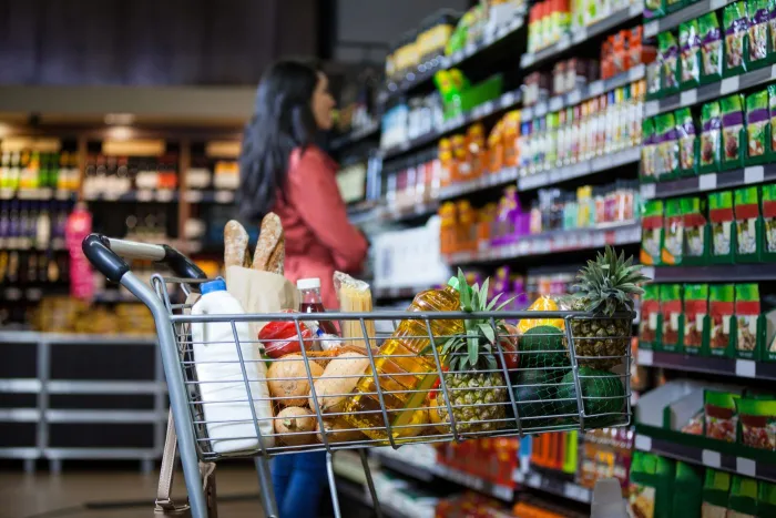 Various groceries in shopping cart in the foreground; a woman reads labels on a shelf of items in the background.