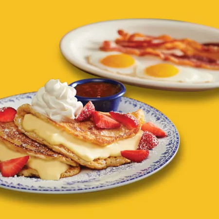foreground: stuffed cheesecake pancake topped with whipped cream and strawberries on plate. background: two eggs, sunny-side-up and strips of bacon on plate