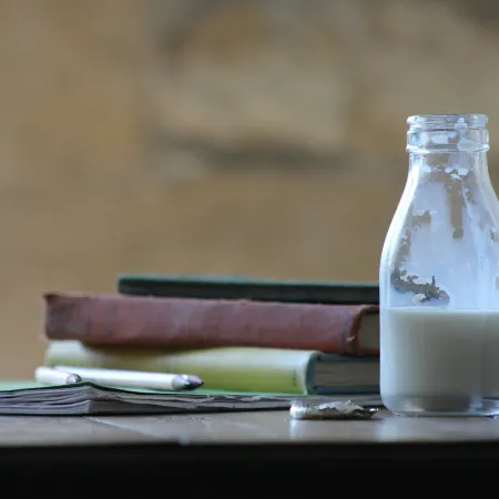 A small bottle of milk on a table beside textbooks and pencils