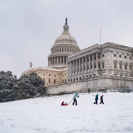 A snowy U.S. Capitol Building with a parent, with their children, pulling a sled uphill 