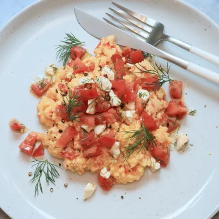 white plate with scrambled eggs, tomatoes and fetta and fork and knife